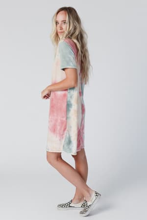 A woman models The Nest On The Daily Oversized Slit Tee Dress - Tie Dye, featuring pastel pink, blue, and cream hues with side slits. She pairs it with black-and-white checkered slip-on shoes and wears her long blonde hair down.