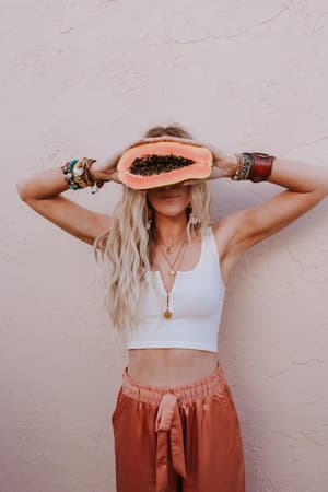 A woman with long blonde hair holds a halved papaya over her eyes against a pink wall, wearing the Hook It Up Ribbed Brami in Ivory, peach pants, layered necklaces, and colorful bracelets.