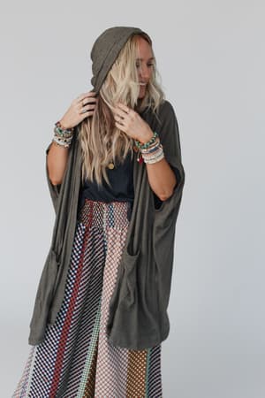 A person with long blonde hair smiles while adjusting the hood of The Nest Boho Essential Hooded Ribbed Sweater in Olive, paired with colorful beaded bracelets, a black top, and a patterned skirt against a plain light background.