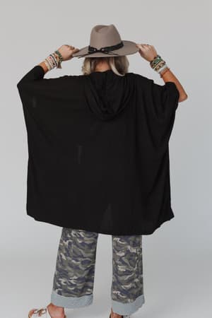 Wearing The Nest Boho Essential Hooded Ribbed Sweater in Black, a person with a wide-brim hat, camouflage pants with striped cuffs, sandals, and multiple bracelets stands against a plain background facing away.