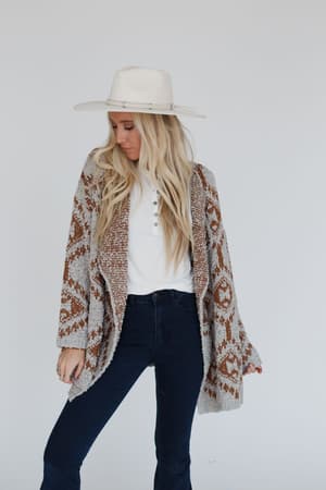 A woman stands against a white background, looking down with one hand by her side and the other holding the Free to Roam Cardigan – Gray, an oversized cardigan with an Aztec-inspired print. She's wearing a white hat, white shirt, and dark jeans.