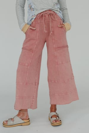 Wearing the Lucky One Wide Leg Pant in Mauve, a striped long-sleeve shirt, and beige sandals, this person stands against a plain background with hands in pockets for an effortless boho vibe.