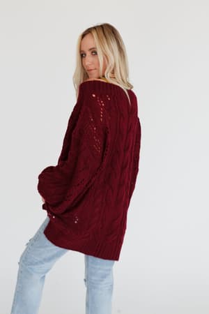 A woman with long blonde hair wears the Golden Hour Bubble Sleeve Sweater Dress in Burgundy, standing slightly turned to the side against a plain white background.