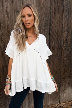 A woman with long blonde hair poses by a wooden fence, wearing the More Than Meets The Eye Tunic Top in ivory with ruffle sleeves, paired with dark jeans and colorful bracelets on both wrists.