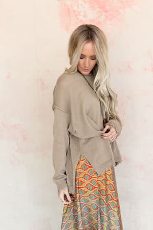 A woman with long blonde hair wears the Billowy Day Sweater - Taupe and a colorful patterned skirt, standing against a pale wall with pink accents, looking down with one hand by her side.