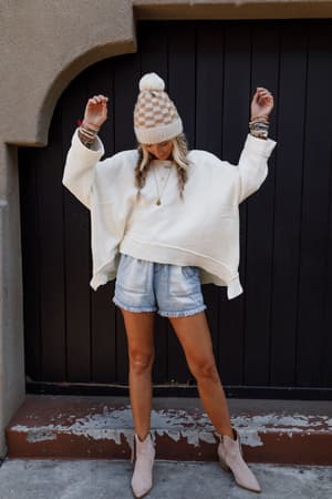 A woman stands with arms raised, wearing the Dana Oversized Diamond Texture Sweater in ivory, a beige pom-pom beanie, light denim shorts, beige ankle boots, and several bracelets in front of a dark wooden door with her head tilted down.