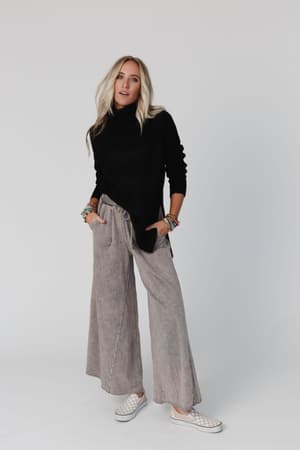 A woman with long blonde hair wears the Danny Sweater in black, paired with loose gray wide-leg pants, bracelets, and checkered slip-on shoes, standing against a plain white background.
