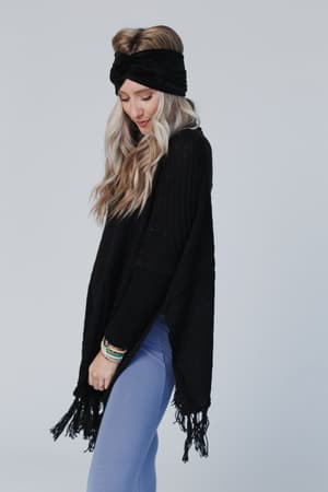 A woman with long, wavy blonde hair wears the Paloma Poncho Sweater in black, styled with a chunky knit black headband and light blue leggings. She stands against a plain light gray background, smiling softly as she looks down.