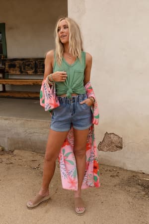 A woman with long blonde hair and bracelets poses casually outdoors in front of a rustic wall, wearing The Nest Shelli Scoop Neck Tank Top in Sage, styled with denim shorts, a long pink floral kimono, and wedge sandals.