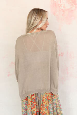 A woman with long blonde hair, seen from behind, wears the Billowy Day Sweater in taupe over a colorful patterned skirt. The background features a light wall with pink and peach watercolor-style patches.
