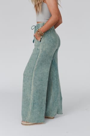 A woman stands in a relaxed pose against a plain background, wearing the Relaxing Robin Wide Leg Pant in New Teal with a drawstring waist and pockets, paired with a cropped sleeveless top and bracelets.