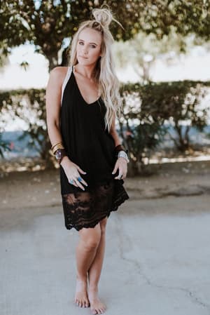 A woman with long blonde hair stands barefoot outdoors on a concrete path, wearing the Prima Lace Hem Slip - Black and several bracelets. Bushes and trees are visible in the background.