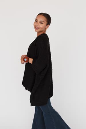 A woman with dark hair tied back smiles and poses sideways in the So Deserving Knit Ruana - Black, paired with flared blue jeans, earrings, and a watch, against a plain white background.