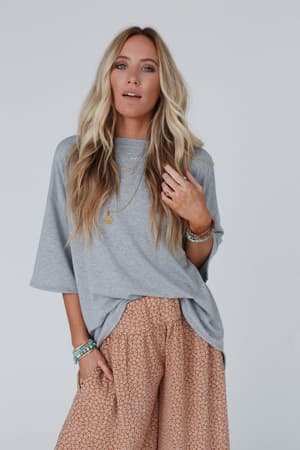 A woman with long, wavy blonde hair wears The Nest Must Have High Low Tee in Heather Gray and patterned peach wide-leg pants, accessorized with layered jewelry for a chic boho look against a light background.