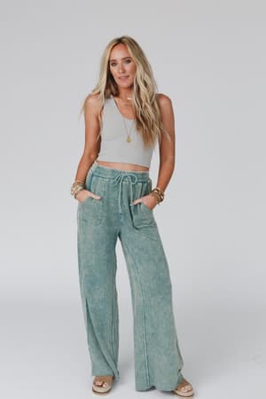 A woman with long blonde hair wears the Relaxing Robin Wide Leg Pant in New Teal with a gray sleeveless top, sandals, and bracelets, standing against a plain light background with her hands in her pockets.