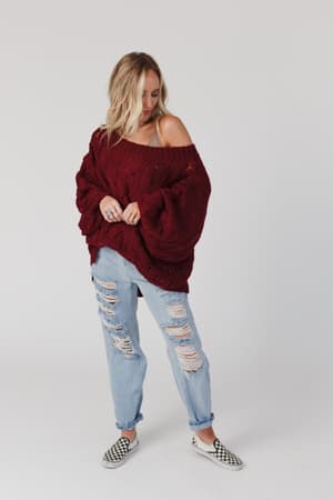 A person wearing the Golden Hour Bubble Sleeve Sweater Dress in burgundy stands against a plain white background, looking down with hands holding the dress.