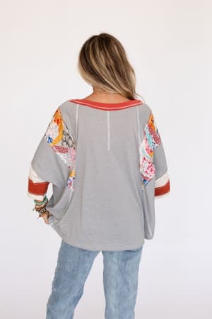 A woman with light brown hair and bracelets wears the Britt Patchwork Top in Heather Gray, featuring an oversized boho fit with colorful patchwork on the 3/4 sleeves and shoulders, paired with light blue jeans against a plain white background.