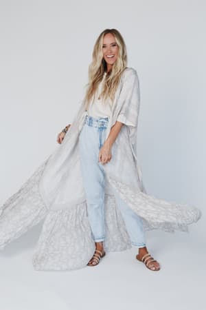 A woman with long blonde hair smiles while wearing the Paisley Tapestry Free Flow Duster Kimono in gray, paired with high-waisted light blue jeans, a white top, and brown sandals against a plain white background.