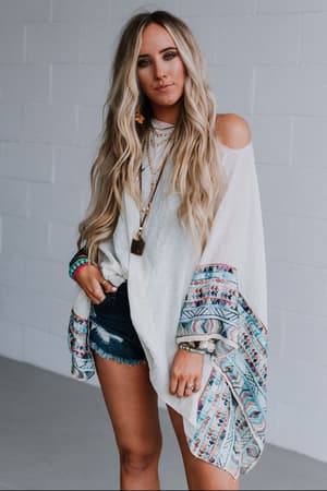 A woman with long wavy blonde hair wears the Fallon Embroidered Sleeve Poncho in ivory, styled with denim shorts, layered necklaces, bracelets, and poses against a white brick wall.