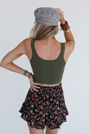A woman stands facing away in a gray hat, bracelets, black floral shorts, and the Hook It Up Ribbed Brami in olive. Her hand touches the back of her head against a plain white background.