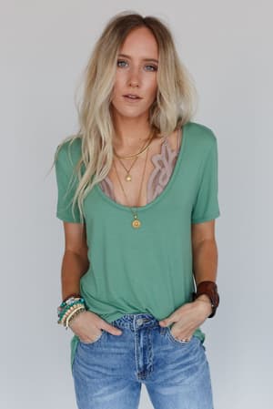 A woman with long blonde hair wears the Scoop Neck Bralette Tee in Sage, styled with layered necklaces, bracelets, and blue jeans. She stands against a plain light gray background with her hands in her pockets.
