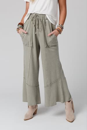 A person wearing a white T-shirt, Feeling Good Wide Leg Pant - Faded Sage, beige ankle boots, and colorful beaded bracelets stands with hands in pockets.