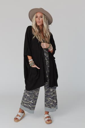 A woman with long blonde hair stands confidently in The Nest Boho Essential Hooded Ribbed Sweater - Black, a wide-brimmed hat, a long black kimono, camouflage wide-leg pants, and colorful bracelets and rings.
