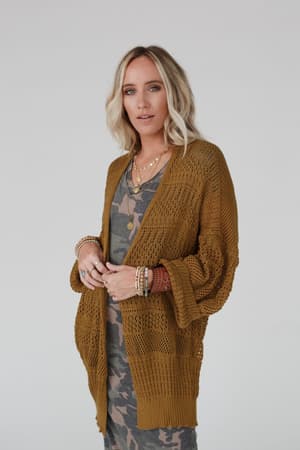 A woman with blonde hair wears the Brenden Knit Netted Cardigan in brown over a green camo print dress, accessorized with layered necklaces and bracelets. She stands against a plain light background.