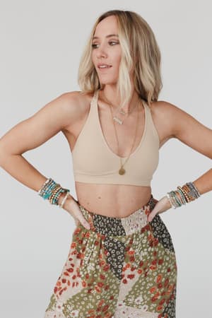 Woman with shoulder-length blonde hair wears the Evermore Seamless Lace Racerback Bralette in beige, a floral skirt, layered necklaces, and beaded bracelets, standing hands on hips and looking to the side against a light background.