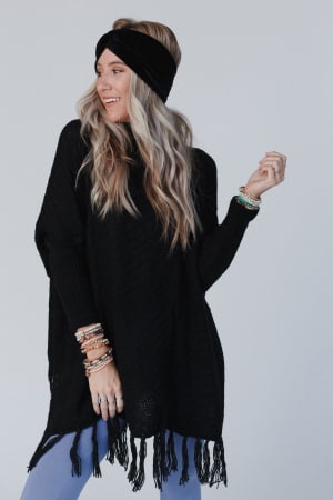 A woman with long, wavy blonde hair smiles and looks to the side against a plain background, wearing the Paloma Poncho Sweater - Black with fringe at the hem, a black headband, blue leggings, and stacked bracelets.