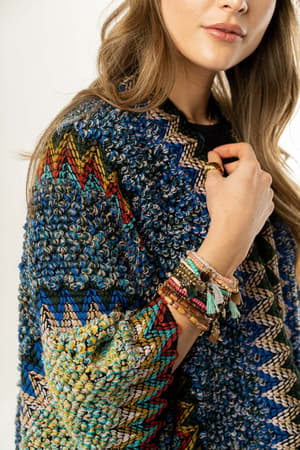 A woman wears the Desert Dreams Crochet Ruana in Cobalt, featuring textured, multicolored zigzag patterns, paired with beaded and tassel bracelets. Her wavy hair falls over her shoulders, and her face is partially visible.