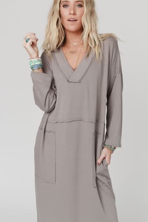 A woman with wavy blonde hair wears The Nest Meadow Long Sleeve Slit Maxi Dress in Coco, a loose-fit style with large front pockets. She accessorizes with layered bracelets and necklaces, standing against a plain light background.