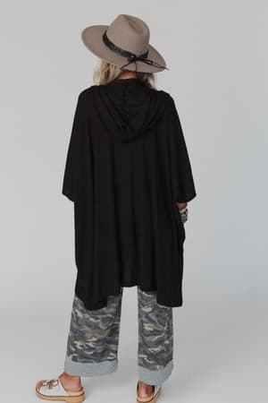 A person faces away from the camera, wearing The Nest Boho Essential Hooded Ribbed Sweater in black, styled with camouflage wide-leg pants with striped cuffs and sandals, against a plain light background.