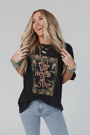 A smiling woman with long blonde hair wears the Share The Love Graphic Tee in charcoal, paired with light blue jeans and colorful bracelets, standing against a light background.