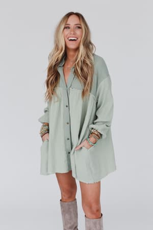 A woman with long, wavy blonde hair smiles in the Sunday Kind Of Love Dress - Sage, a loose oversized light green dress with pockets. She accessorizes with bracelets and knee-high taupe boots, standing against a plain light background.