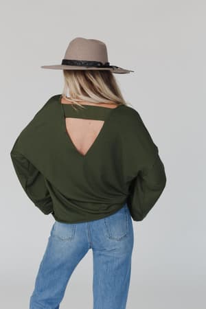 A person wearing a wide-brimmed beige hat, light blue jeans, and The Nest Love It V Neckline Sweater Top in olive with a V-shaped back cutout stands facing away, hands on hips, against a plain light background.