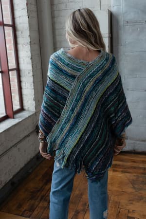 A woman with long blonde hair stands indoors, facing away, wearing the Madeleine Poncho - Blue, which features colorful diagonal stripes in blue, green, and gray. She pairs it with blue jeans beside a brick wall and large window.