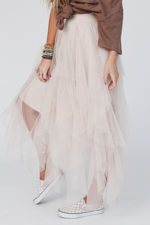 A person wears the Boho Ballerina Skirt - Nude with a brown rolled-sleeve top, multiple bracelets, and checkered slip-on sneakers, standing against a plain white background.