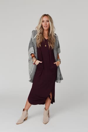 A woman with long blonde hair wears the Call It Comfort Tee Dress in Plum with a gray patterned shawl, multiple bracelets, layered necklaces, and beige ankle boots, standing against a plain white background with her hands in her pockets.