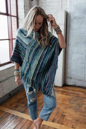 A woman wears the Madeleine Poncho - Blue with distressed jeans and beaded bracelets, standing barefoot on a wooden floor in a bright industrial-style room with brick and white walls.