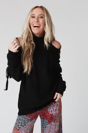 A smiling woman with long blonde hair wears the Roxy Criss Cross Tie Shoulder Sweater in black, standing against a plain light background.