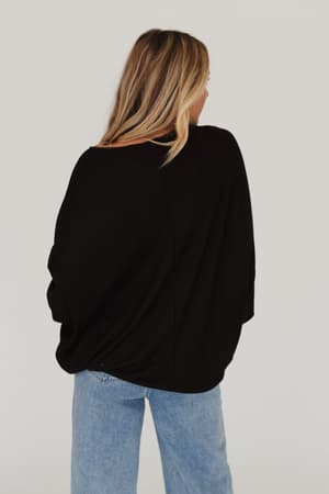 A woman with light brown hair and blonde highlights stands facing away, wearing the Fly With Me Batwing Sleeve Knit Sweater in black and light blue jeans against a plain white background.