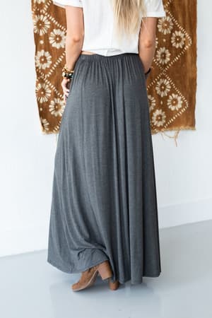 A woman with long blonde hair wears The Perfect Pocketed Maxi Skirt in charcoal and a white shirt, standing with her back to the camera. She pairs it with brown ankle boots and bracelets, while a patterned brown cloth hangs on the wall.