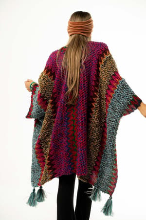 A woman with long hair, wearing a headband and black pants, is shown from the back in the Desert Dreams Crochet Ruana - Magenta—a vibrant knit poncho with tassels that adds boho style to any closet.