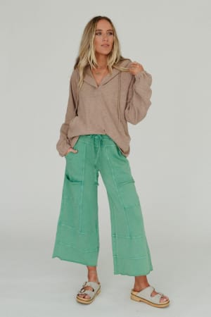A woman with long, wavy blonde hair poses against a plain background in boho style, wearing the Lucky One Wide Leg Pant - Green with patchwork details, a loose beige top, and light-colored sandals.