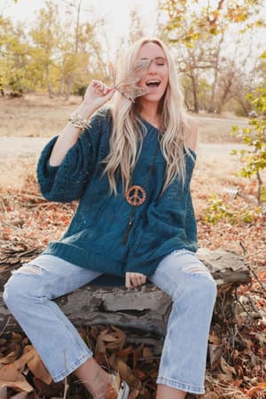 A smiling woman with long blonde hair sits outdoors on a log, wearing the Golden Hour Bubble Sleeve Sweater Dress in teal. She holds a leaf near her face, surrounded by scattered autumn leaves and accessorized with a peace sign necklace.