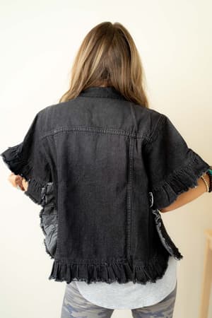 A person with long hair faces away from the camera, wearing the Scottie Ruffle Denim Jacket - Black over a light shirt and patterned leggings. The jacket features a cropped hem and frayed edges. The background is plain and light-colored.