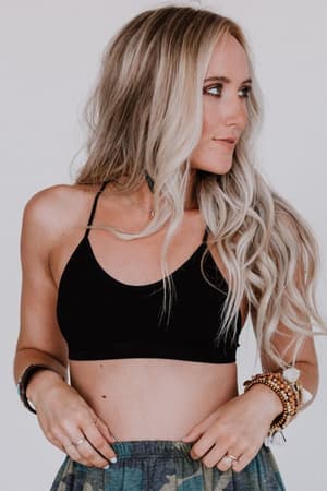 A woman with long, wavy blonde hair looks to the side. She wears the Tattoo Bralette - Black as a crop top, paired with a patterned skirt and several bracelets on her left wrist. The background is plain and light-colored.