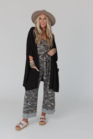 A woman wears The Nest Boho Essential Hooded Ribbed Sweater in black over a camouflage jumpsuit, styled with a wide-brimmed hat, white sandals, and colorful bracelets, smiling against a plain light background.