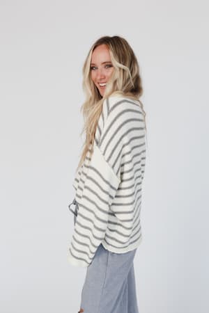 A woman with long blonde hair smiles over her shoulder, wearing the All My Stripes Oversized Sweater in Ivory Gray and light blue pants, standing against a plain white background.